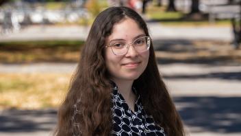 A person with glasses and a black and white patterned shirt standing outside in front of a shaded pathway