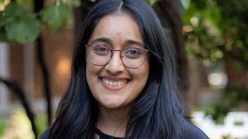 A person with dark hair and glasses standing in front of a tree