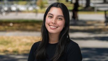 A person with long dark hair in a black shirt standing in front of a row of trees