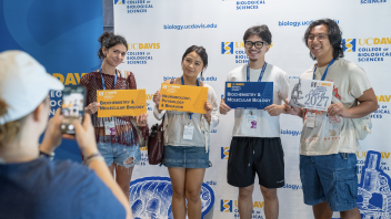 Four smiling students pose for a photo, holding props, while a CBS faculty member takes the picture on a smartphone.