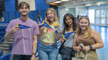 Four students smile at the camera, posing and holding up items that were distributed at New Student Orientation.