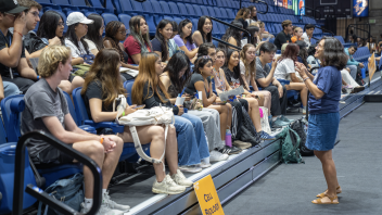 A UC Davis professor wearing a blue shirt addresses a large cohort of new students sitting on bleachers.