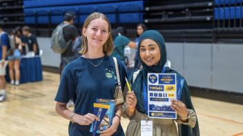 Two students smile for a picture, holding up a sheet of stickers, a small pendant, and other materials received at the event.