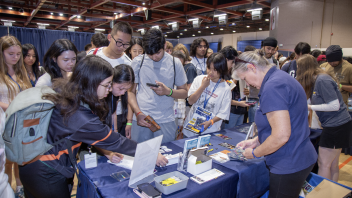 Students gather around a booth at an event, engaging with materials and asking questions to a staff member.