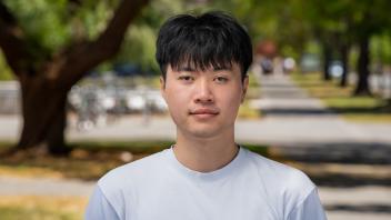 A person standing outside in a grey t-shirt with a row of trees and a pathway behind them.