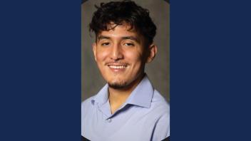 A person with short, curly hair and facial hair is smiling in this portrait. They are wearing a light blue shirt, and the background is a neutral, blurred setting.