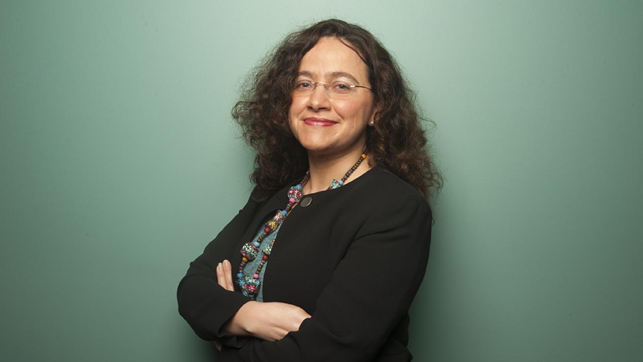Smiling woman with curly hair and glasses, arms crossed, black blazer, teal background