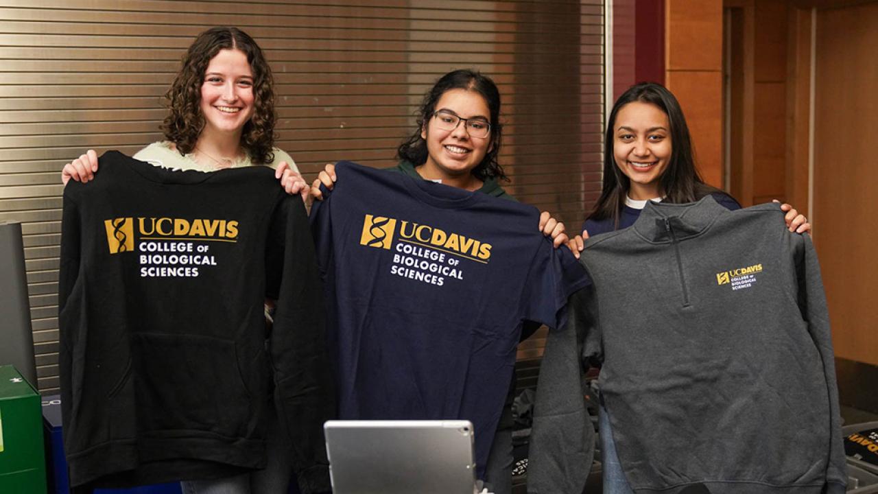 Three smiling young women stand behind a table displaying UC Davis College of Biological Sciences merchandise, holding up sweatshirts and T-shirts with the college logo. They are indoors, and the table in front of them is covered with neatly folded apparel.