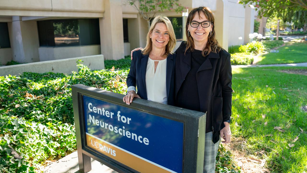 Two women standing in front of the Center for Neuroscience building next to the blue and gold Center for Neuroscience sign