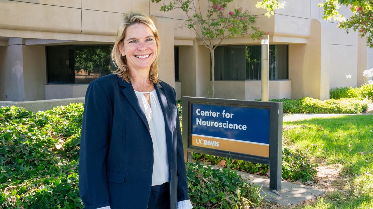 Smiling woman in blazer stands outside UC Davis Center for Neuroscience building beside center’s sign.