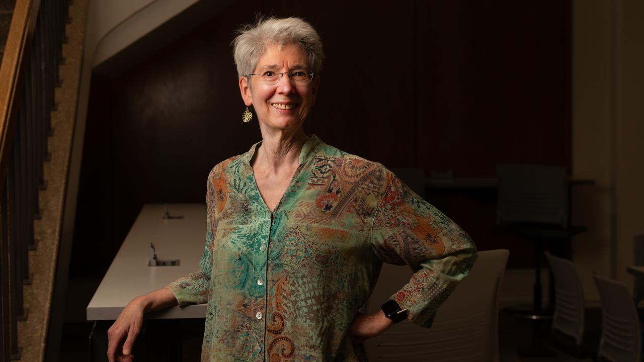 Smiling person with short gray hair, glasses, and patterned shirt standing indoors with hand on hip.