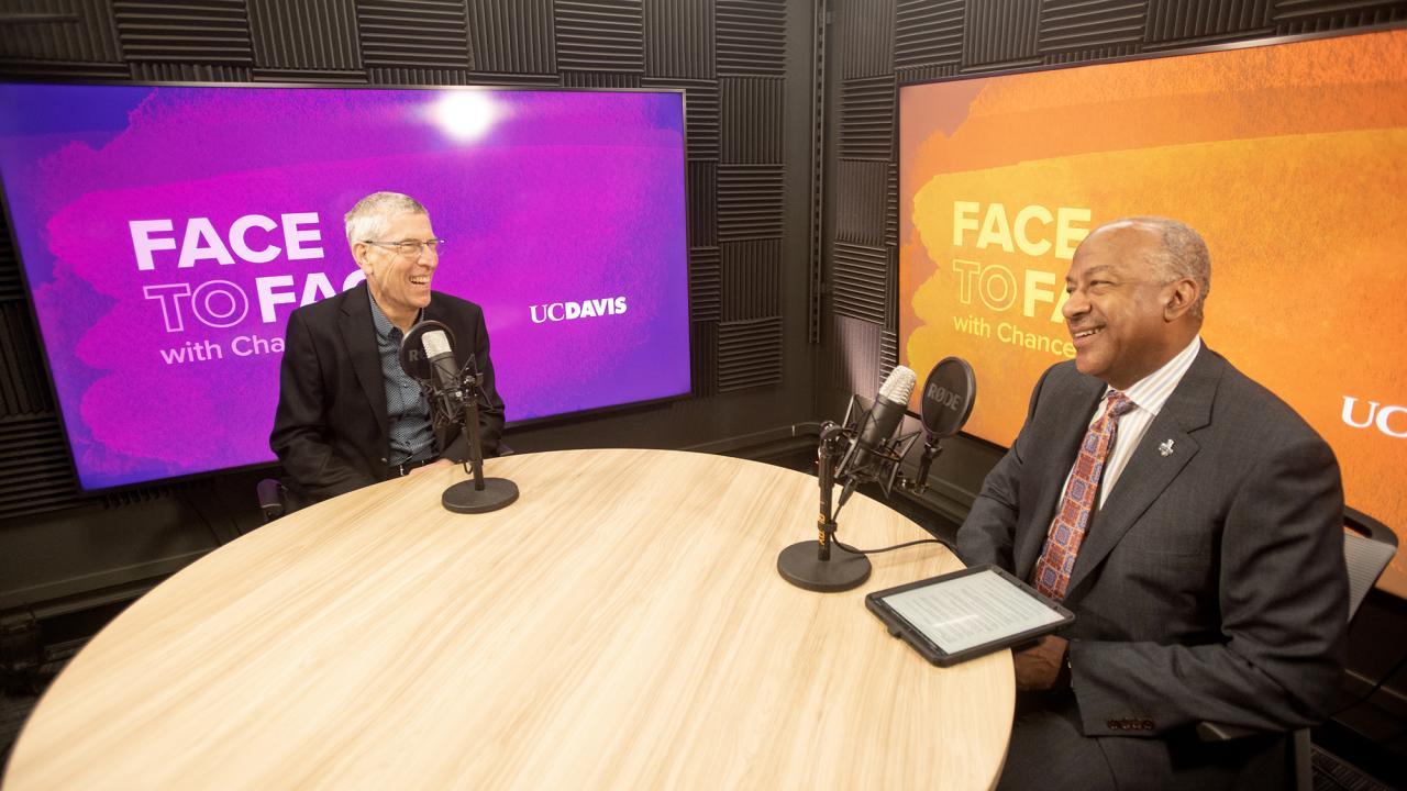Two men sit at a round table with microphones, smiling and engaging in conversation.