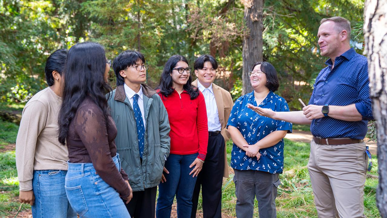 Instructor gestures while talking with smiling students gathered in the arboretum surrounded by trees and greenery.