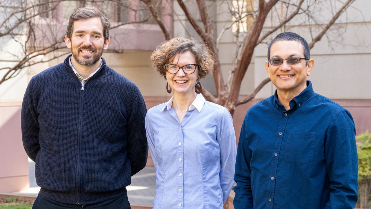 Three smiling individuals stand outdoors in front of a beige building with leafless trees, dressed in professional attire.