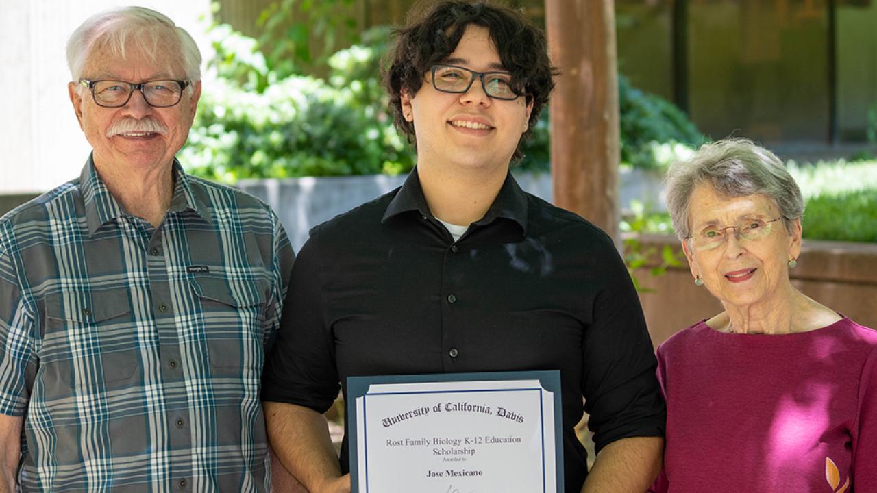 A group of three people standing beneath a tree. The middle person is holding a certificate. 