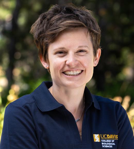 A woman with short brown hair and a friendly smile wears a navy UC Davis College of Biological Sciences polo shirt, standing outdoors with a blurred green background.