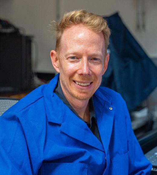 Smiling man with blond hair wearing a blue lab coat in a workshop