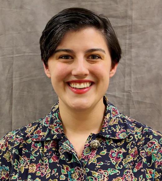 Smiling person with short dark hair wearing a floral shirt against a gray backdrop.