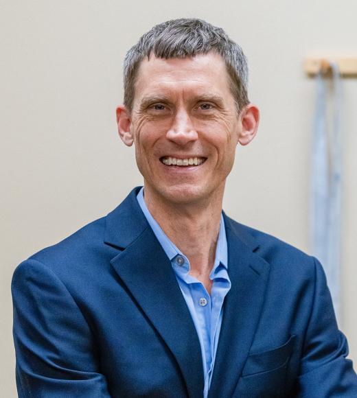 Portrait of a middle-aged man with short, graying hair, wearing a navy blue blazer over a light blue dress shirt. He is smiling warmly and looking directly at the camera, with a neutral beige background featuring a blurred object on the right side.