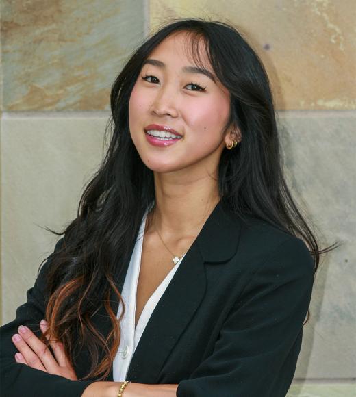 Smiling young woman with long dark hair in black blazer, arms crossed, stone wall background