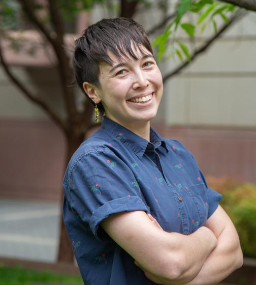 Smiling person with short dark hair in blue patterned shirt, arms crossed outdoors