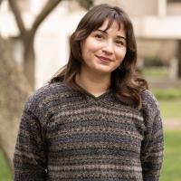 Azucena Virgen, a student with medium-length brown hair, wearing a multicolored knit sweater, smiles while standing outdoors with blurred trees and buildings in the background.