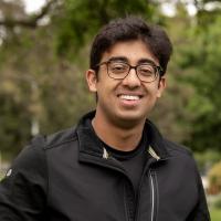 Smiling young man wearing glasses and a black jacket, standing outdoors with trees in the background.