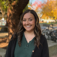 A smiling woman with wavy brown hair, wearing a green shirt and black cardigan, stands outdoors near autumn trees and bicycles.