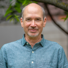 Middle-aged man with short beard, teal button shirt, slight smile, outdoor portrait.