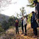 A group of five people stands on a dirt trail in a sunlit forested canyon, listening to one person speak while the others observe and engage. Sunlight filters through trees, and the terrain is dry and hilly with patches of green vegetation.