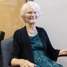 Elderly smiling woman with white hair seated in an armchair wearing teal dress and black cardigan.