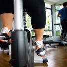 Close-up of a person’s legs pedaling on a stationary exercise bike in a gym, with another individual running on a treadmill in the background.