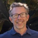 A headshot of a man with short grey-black hair, glasses and a blue shirt standing in front of a palm tree.