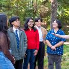 Instructor gestures while talking with smiling students gathered in the arboretum surrounded by trees and greenery.