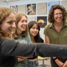 Four students smile and laugh while looking at a screen in a classroom or lab setting. One student in the foreground points at the screen, while the others stand closely together. The room has colorful photos of marine life on the wall in the background.