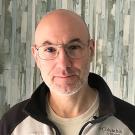 A middle-aged man with a bald head and short gray beard is standing in front of a patterned wall with vertical brushstroke designs in shades of gray, blue, and white. He is wearing large, wire-rimmed glasses and a Columbia Titanium fleece jacket over a beige shirt. He has a neutral expression and is looking directly at the camera.