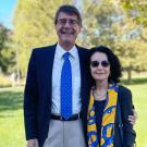 A man and woman stand together outdoors on a sunny day, both smiling slightly. The man wears a dark suit with a white shirt and a blue tie, while the woman wears a dark outfit with a colorful yellow and blue scarf. They appear relaxed, with trees and green grass in the background.