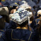 A close-up of a decorated graduation cap adorned with blue flowers, pearls, and a ribbon, surrounded by other graduates in blue and gold academic regalia.