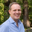 Smiling man in blue patterned shirt, outdoor portrait with trees and dappled sunlight
