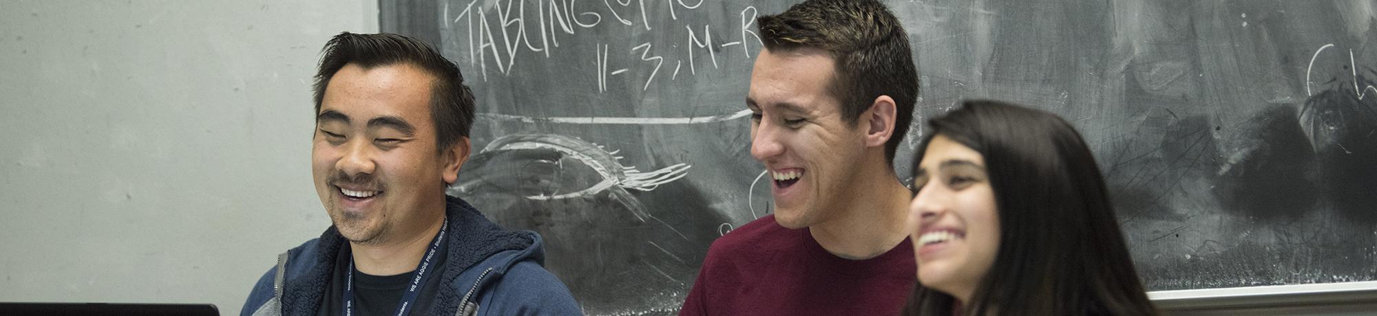 Three students smiling and laughing in a classroom, sitting in front of a chalkboard with writing and diagrams in the background.