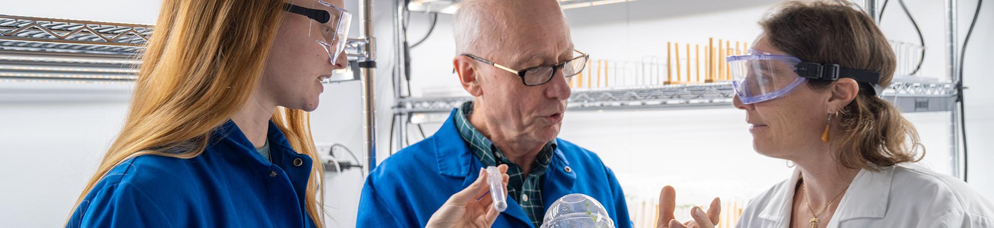 Three people in lab goggles and lab coats examine a small plant sample together in a bright lab setting