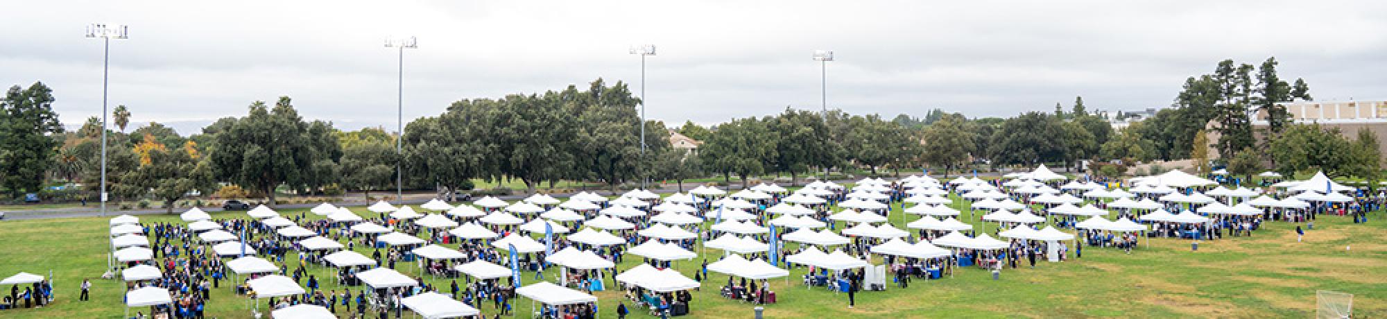 A wide aerial view of a large outdoor event, featuring rows of white canopy tents set up on a grassy field. Attendees can be seen walking between the tents, surrounded by trees and tall light poles in the background.