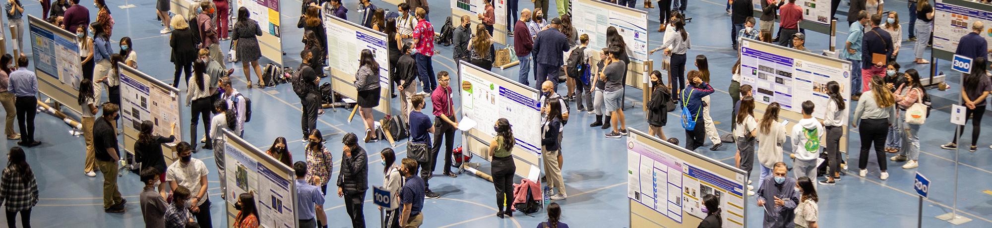 Large indoor research poster session with attendees walking between rows of posters, engaging in discussions. The room is filled with presenters, viewers, and scientific displays on a blue floor.
